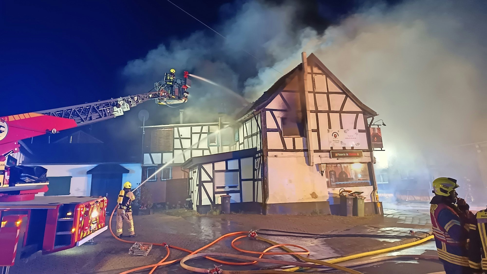 Von einer Drehleiter und aus einem Schlauch spritzen Feuerwehrleute Wasser auf ein qualmendes Fachwerkhaus.