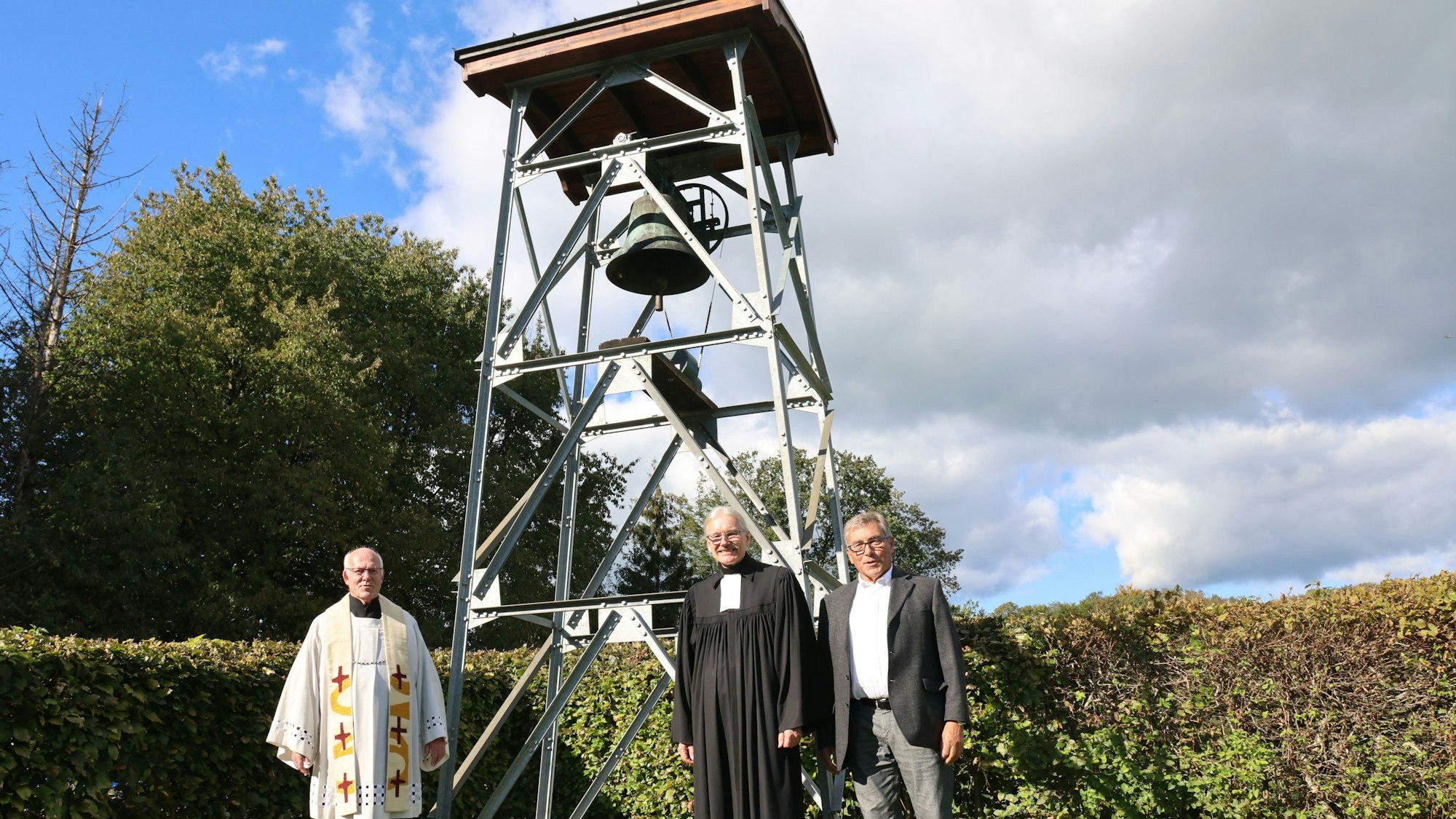 Drei Männer, darunter zwei Geistliche, stehen vor einem neuen Glockenturm.