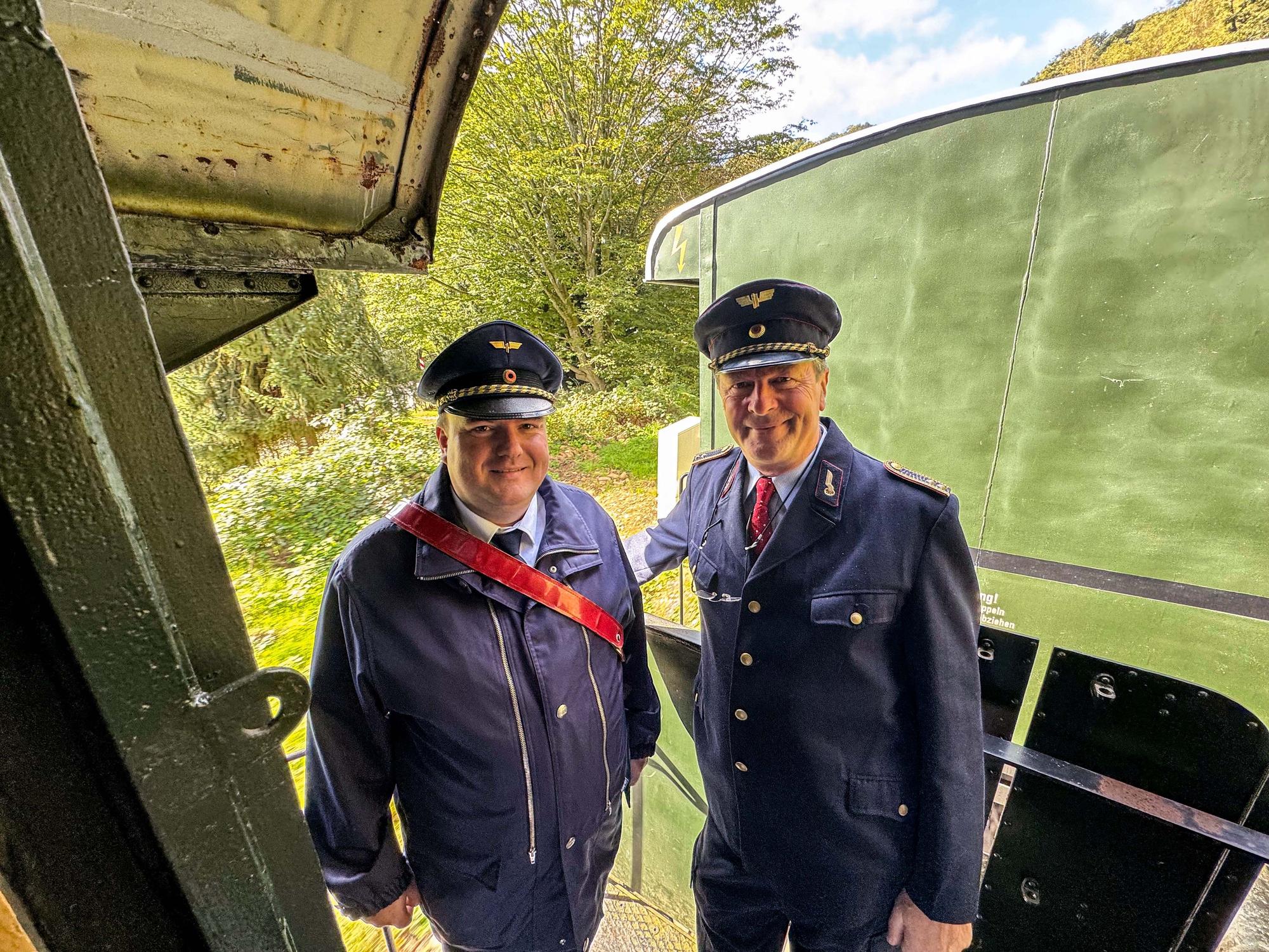 Zwei Männer in Eisenbahn-Uniform stehen während der Fahrt auf der Plattform eines Waggons.