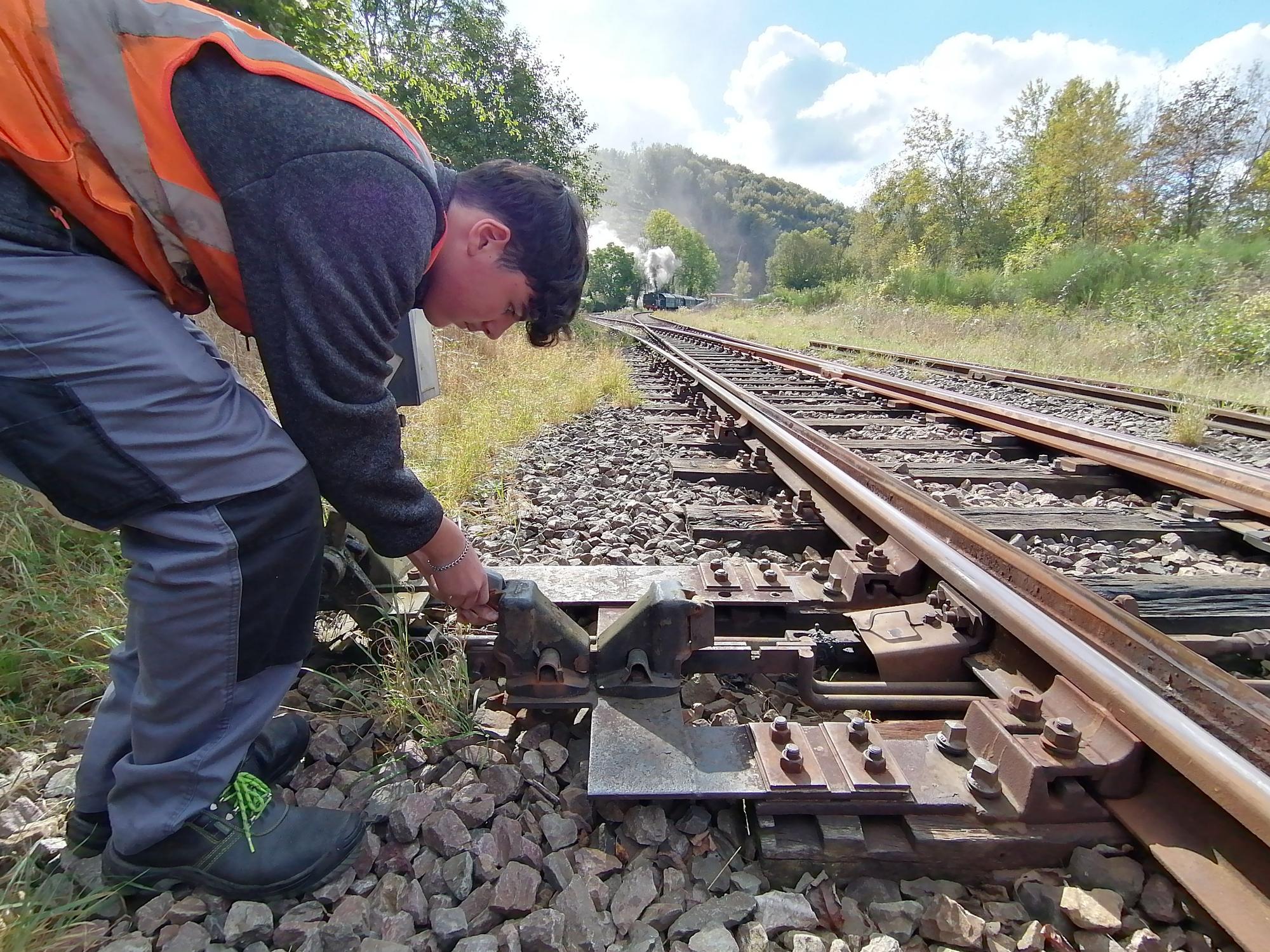 Ein junger Mann mit Warnweste steht über dem Hebel einer Weiche an einem Bahngleis gebeugt.