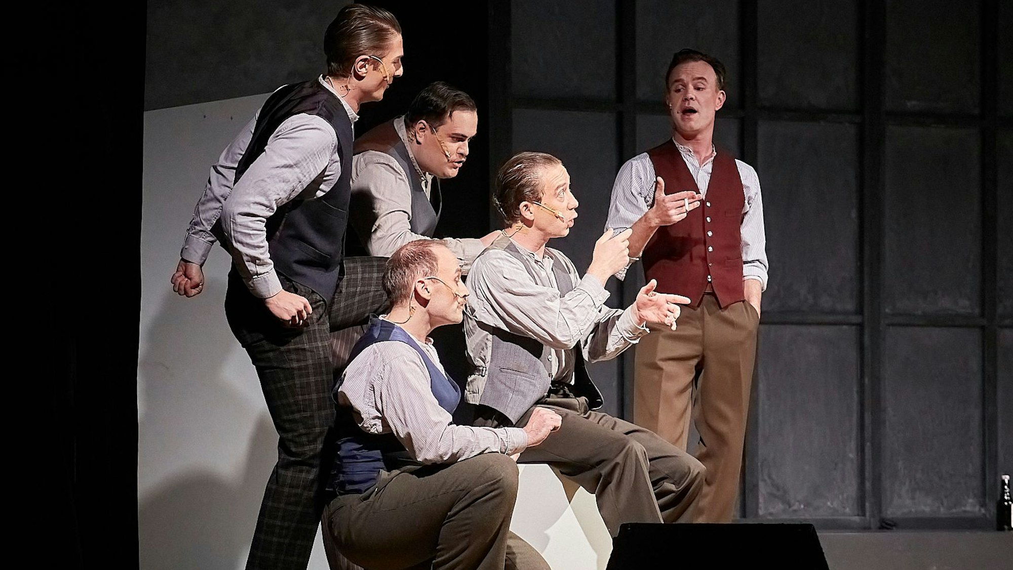 Eine Gruppe von fünf Männern, die im Stil der 1930er Jahre gekleidet sind, stehen gestikulierend auf der Bühne.