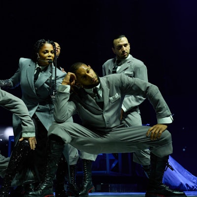 Janet Jackson posiert im stilisierten Business-Outfit inmitten ihrer Tänzer.