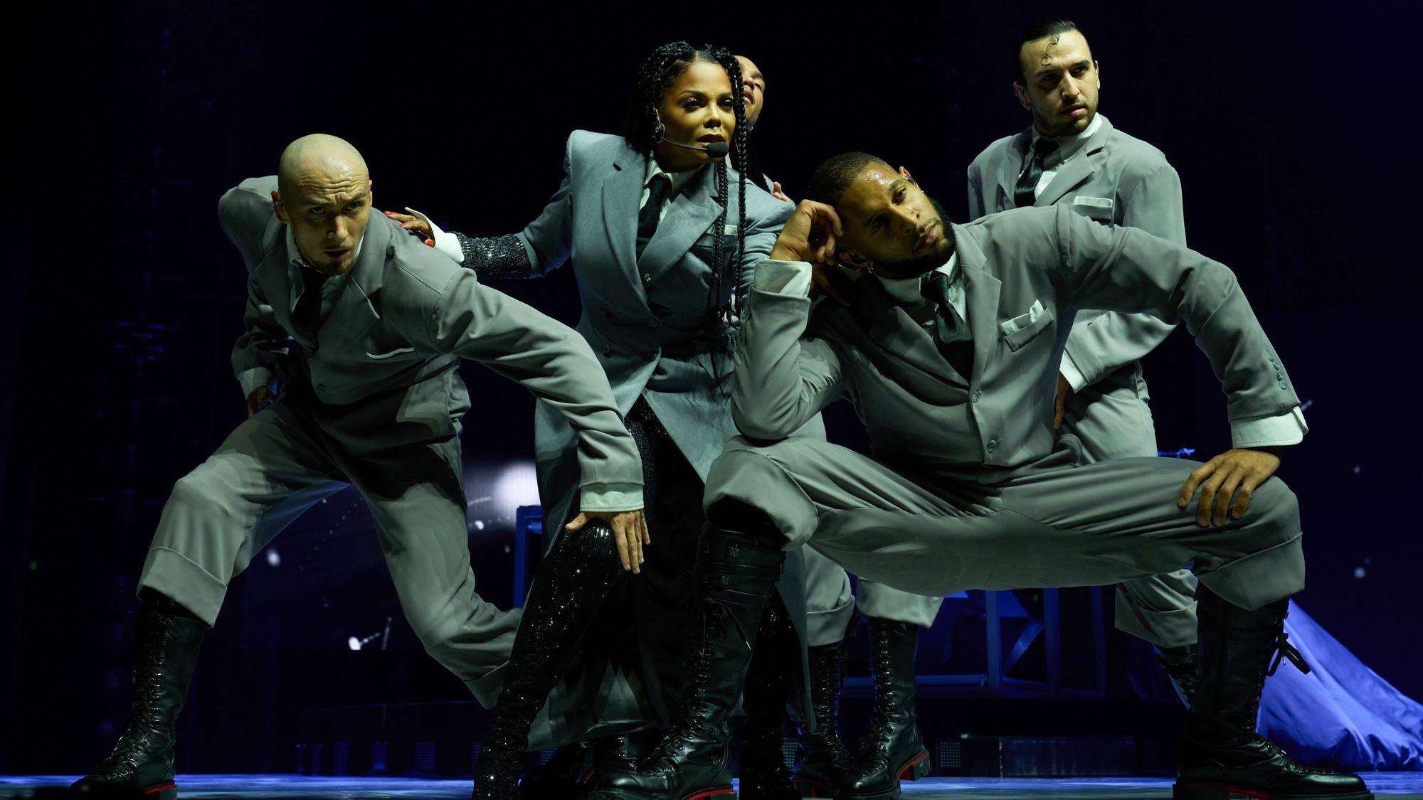 Janet Jackson posiert im stilisierten Business-Outfit inmitten ihrer Tänzer.