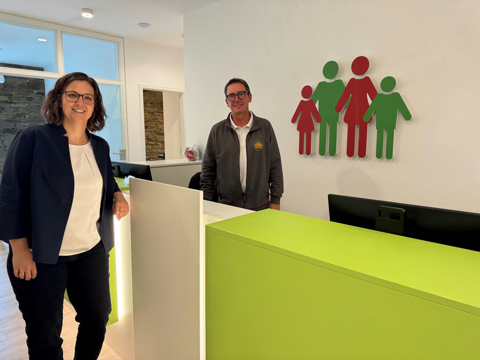 Ptofessorin Dr. Beate Müller, Leiterin des Instituts für Allgemeinmedizin an der Uni Köln und Christian Franchy, Hausarzt in Lindlar, stehen in einem hellen Raum an einer Empfangstheke.