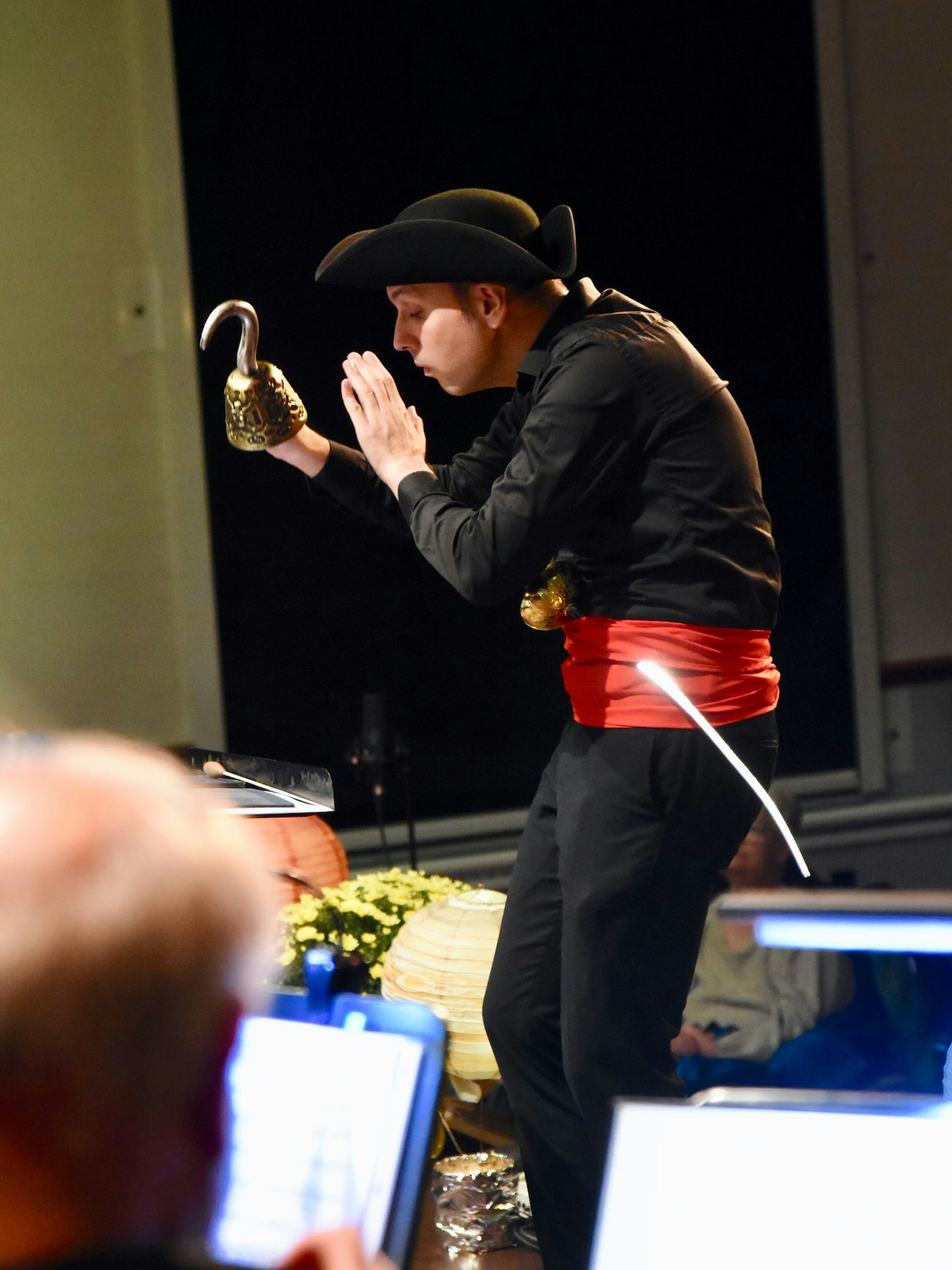 Ein Mann in Schwarz dirigiert ein Orchester mit Hakenhand, Säbel und Dreispitz.