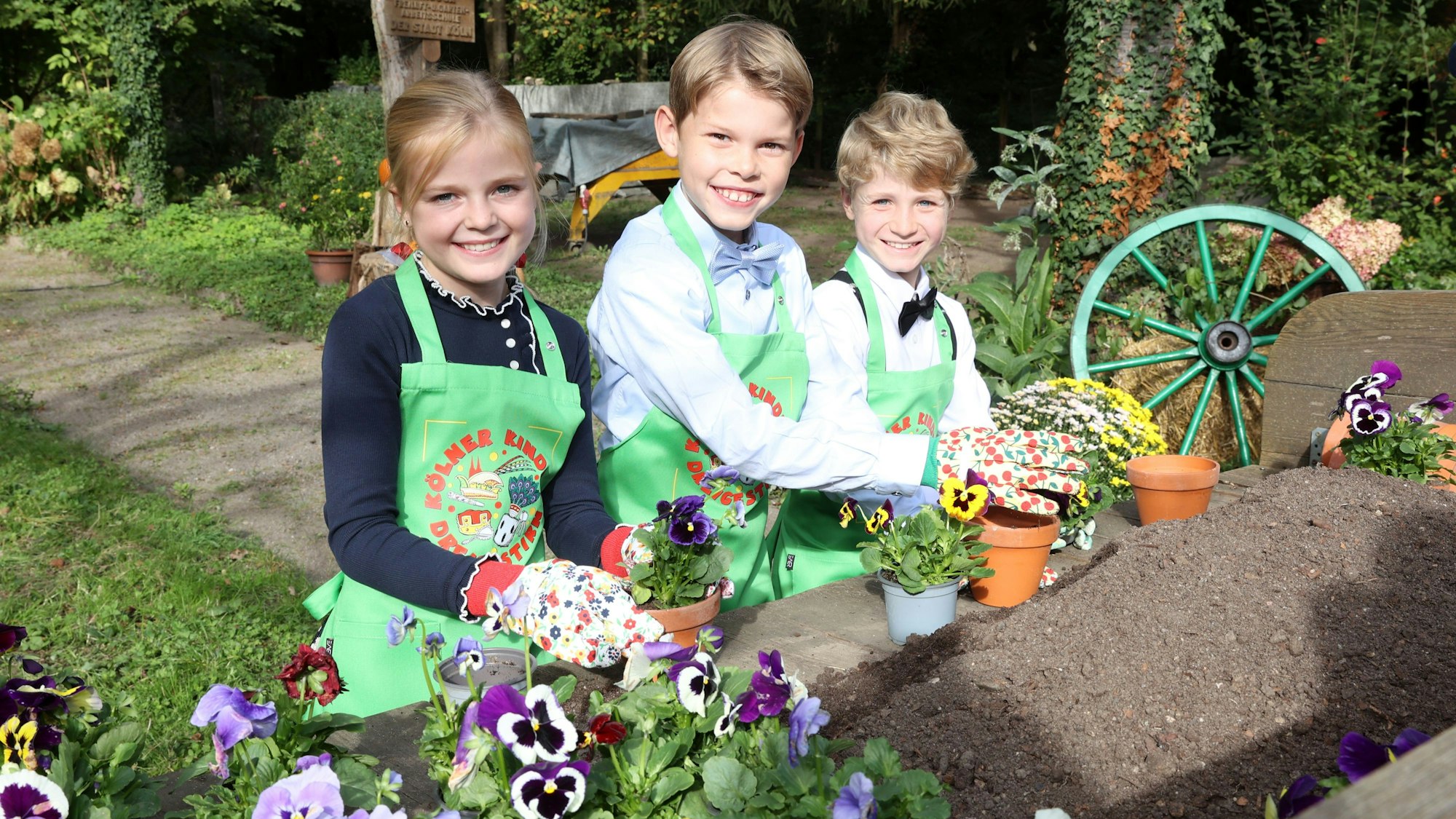 Zwei Jungen und ein Mädchen mit Gartenschürzen und -handschuhen beim Einpflanzen von Blumen.