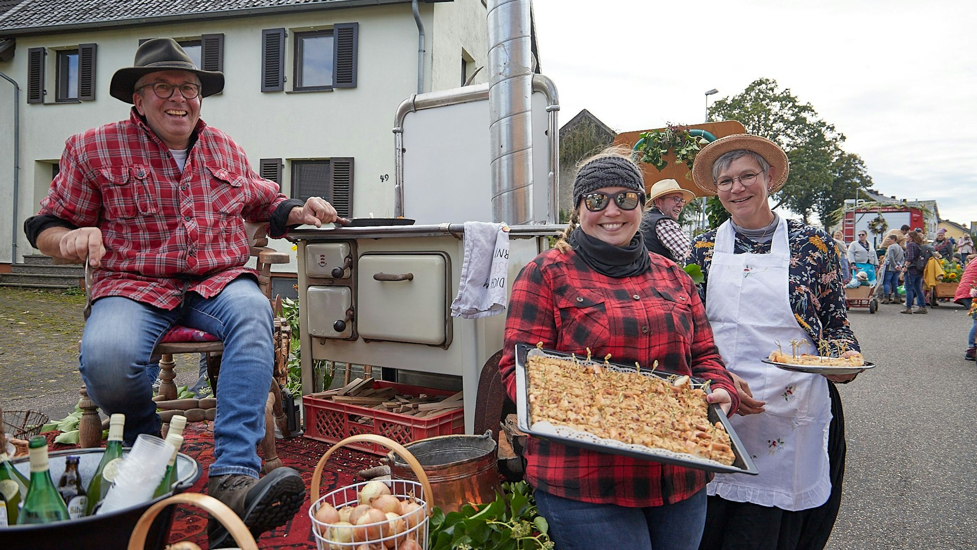 Ein Mann sitzt auf einem Stuhl, der auf einem Wagen steht, eine Frau präsentiert ein Blech mit Zwiebelkuchen.