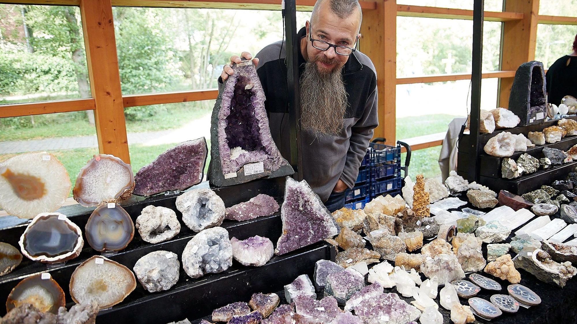 Ein bärtiger Mann steht hinter seinem Verkaufsstand, an dem er zahlreiche Mineralien anbietet.
