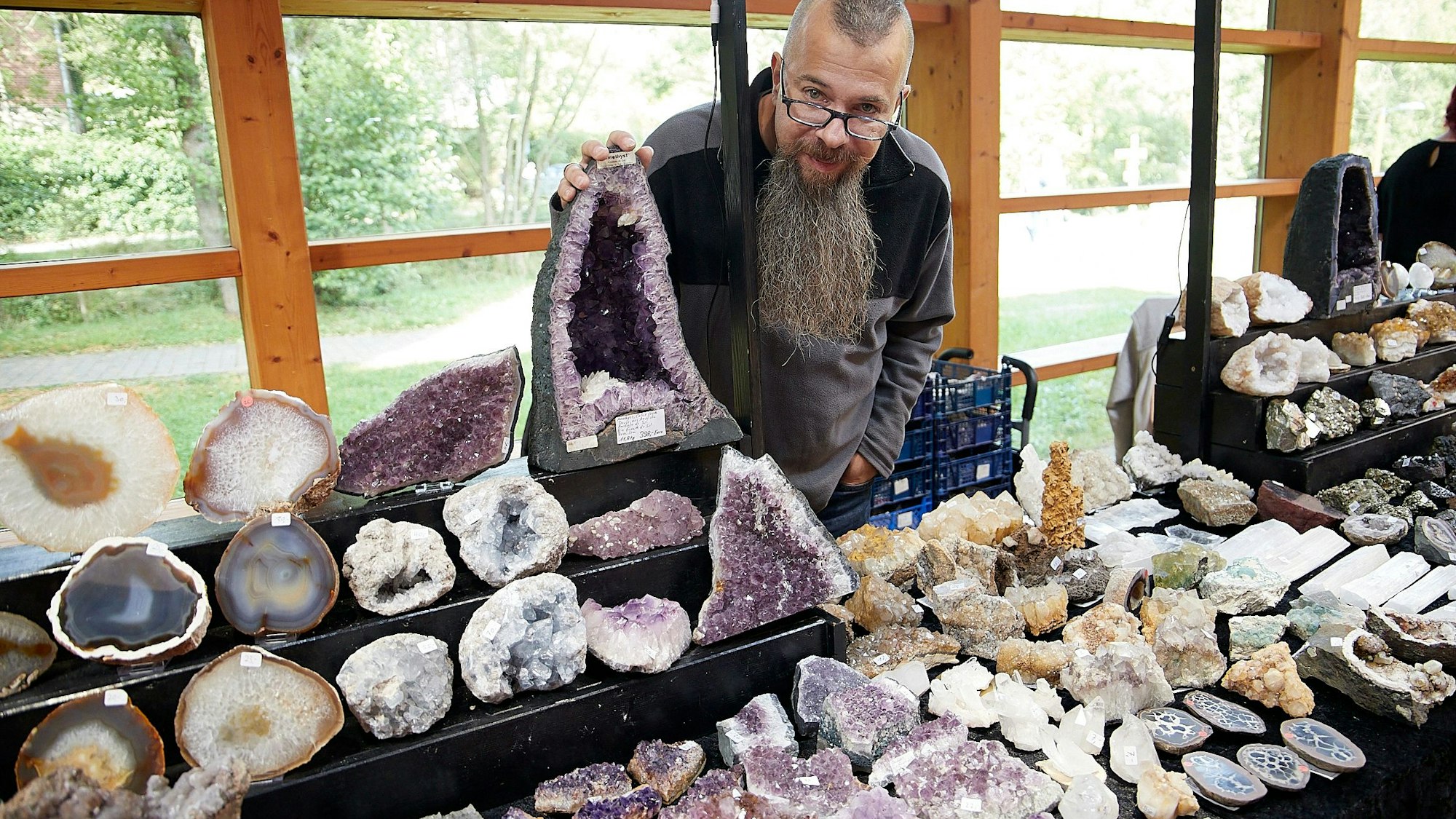 Ein bärtiger Mann steht hinter seinem Verkaufsstand, an dem er zahlreiche Mineralien anbietet.