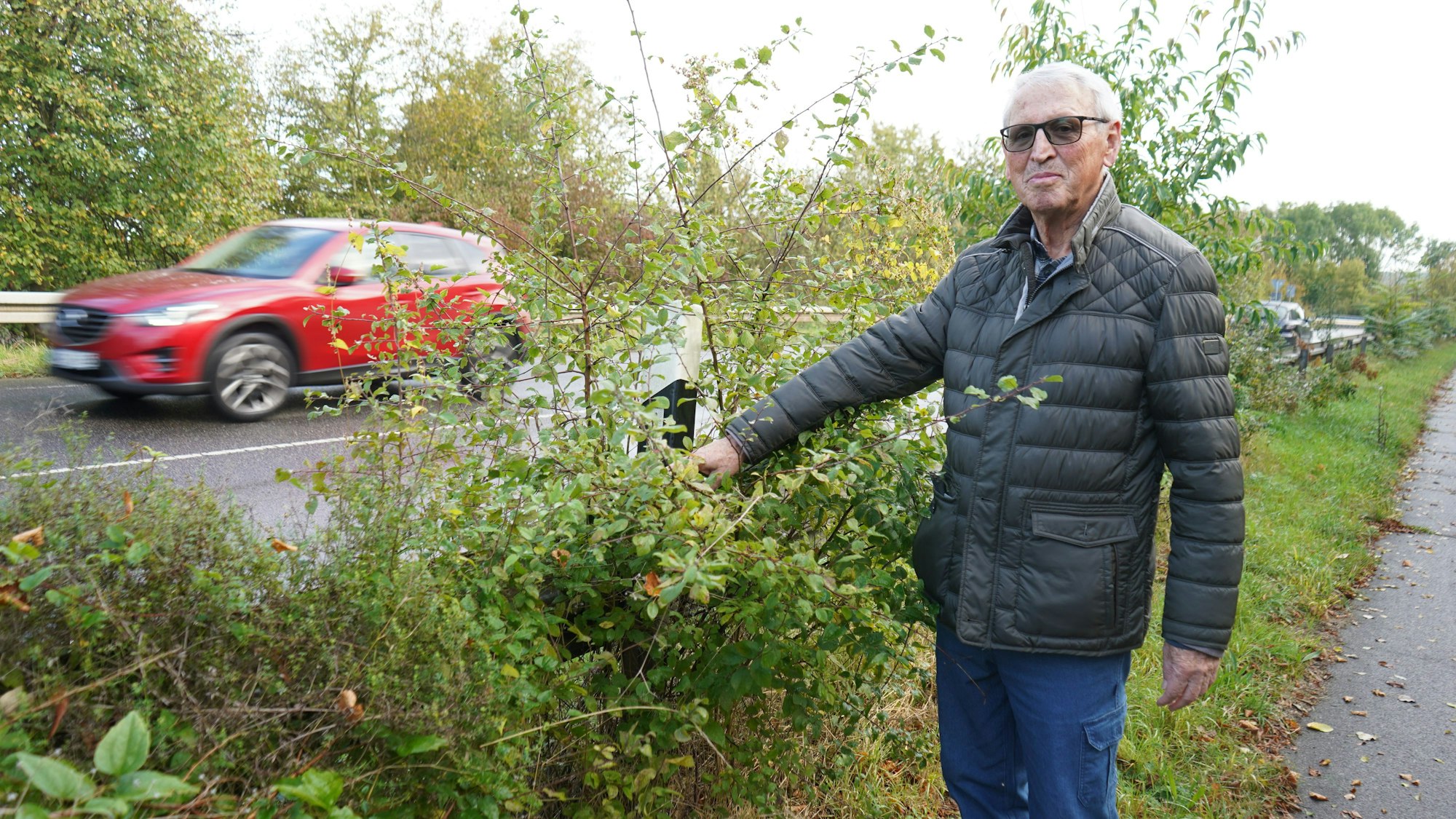 Das Bild zeigt einen Mann, der auf einen Leitpfosten zeigt, der von einem Strauch zugewuchert ist.