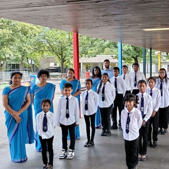 Auf dem Bild sind die Schülerinnen und Schüler zu sehen, die sich in drei Reihen aufgestellt haben, bevor es in die Klassen geht. Sie tragen Schuluniformen. Links und rechts neben ihnen haben sich die Lehrerinnen und Lehrer aufgestellt. Die Lehrerinnen tragen blaue Sahrees.