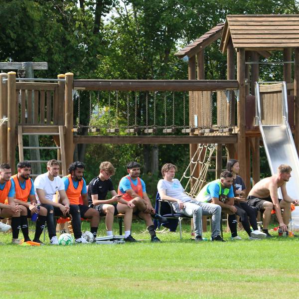 Das Bild zeigt eine Reservebank mit Fußballspielern. Im Hintergrund ist ein Spielplatz zu erkennen.