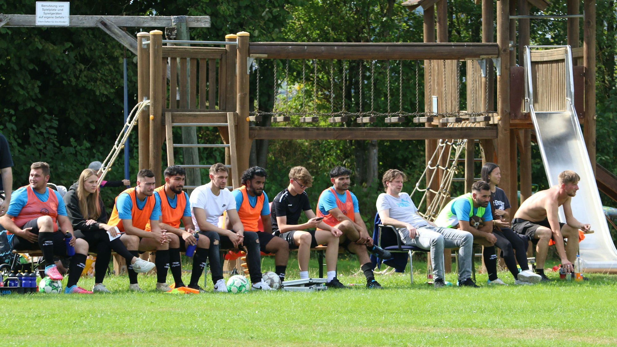 Das Bild zeigt eine Reservebank mit Fußballspielern. Im Hintergrund ist ein Spielplatz zu erkennen.