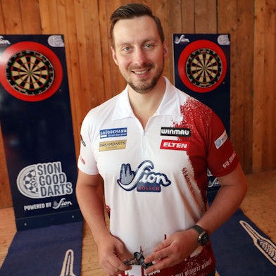 Zu sehen ist der Dartspieler Florian Hempel in der Vorderansicht. Links und rechts hinter ihm sind Dartscheiben zu sehen.