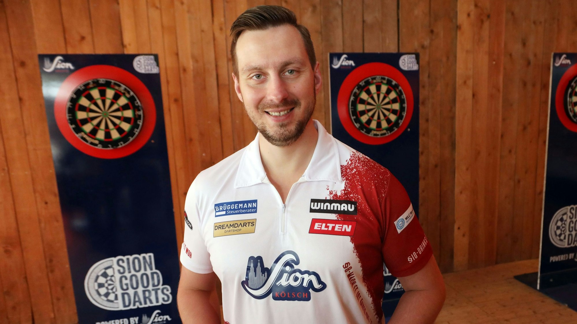 Zu sehen ist der Dartspieler Florian Hempel in der Vorderansicht. Links und rechts hinter ihm sind Dartscheiben zu sehen.