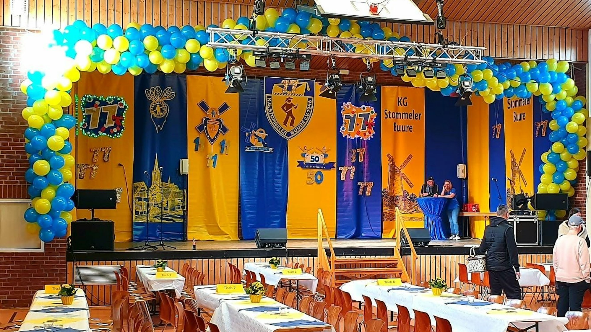 Blick in eine Veranstaltungshalle mit einer Bühne, davor stehen Tische.