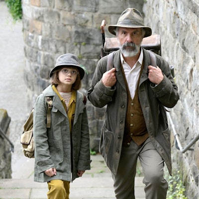 Auf dem Bild sind Christoph Maria Herbst und Yuna Bennett in ihren Rollen als gemeinsame Buchspazierer zu sehen. Sie gehen eine steinerne Treppe hoch. Die beiden tragen Hüte und Jacken.