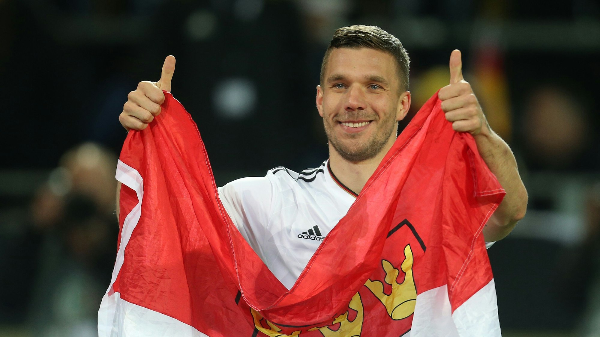 Lukas Podolski verabschiedet sich nach einem Länderspiel von den Fans und hält dabei eine Kölner Fahne.
