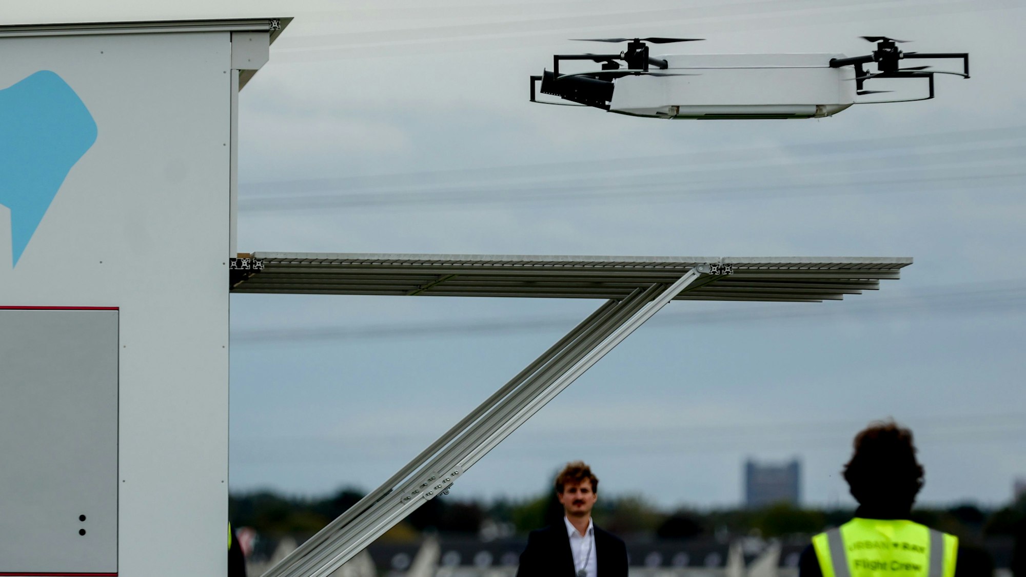 Zu sehen ist eine Drohne im Landeanflug und zwei Personen.