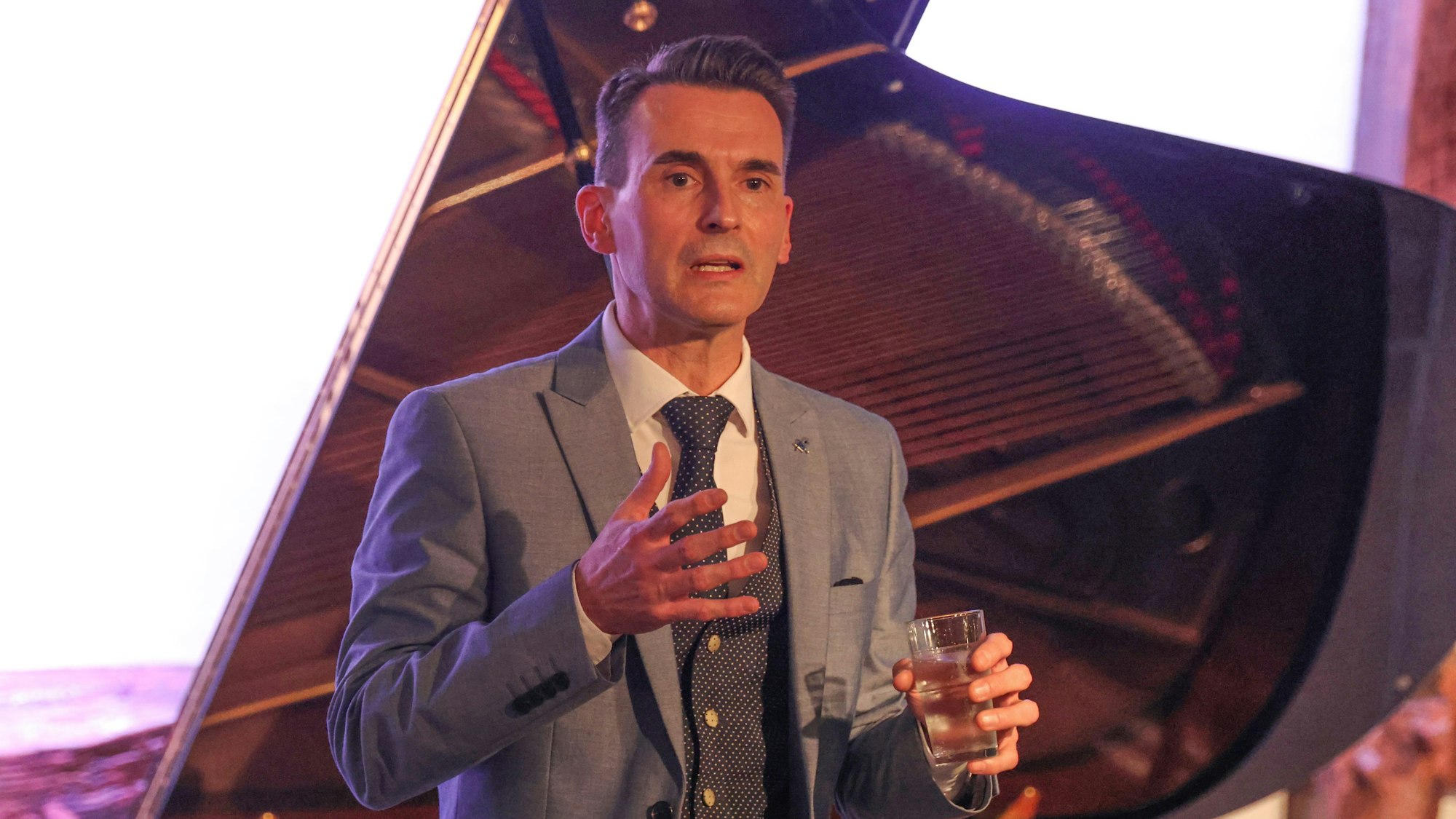 Ein Mann im grauen Anzug spricht vor einem Flügel.