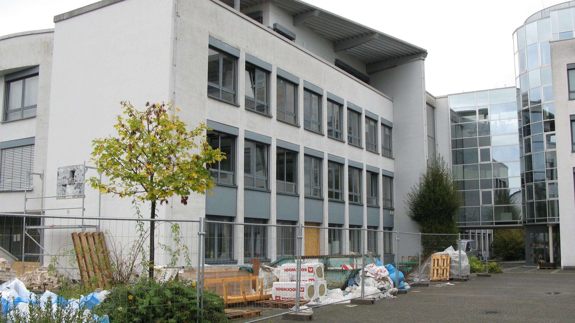 Ein hellgraues, vierstöckiges Bürogebäude mit großer Fensterfront am Zugang und einem Bauzaun und Baumaterialien davor.