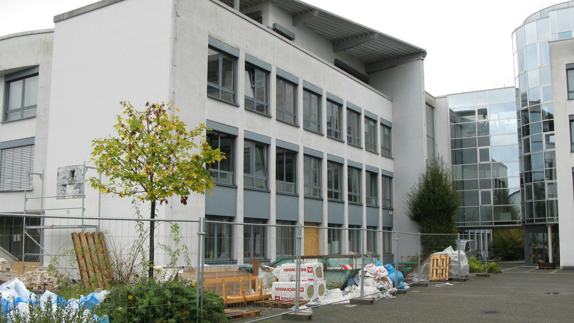 Ein hellgraues, vierstöckiges Bürogebäude mit großer Fensterfront am Zugang und einem Bauzaun und Baumaterialien davor.