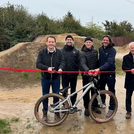 Fünf Männer lächeln in die Kamera, drei sind dabei ein rotes Band zu durchtrennen, vor ihnen steht ein Mountainbike.