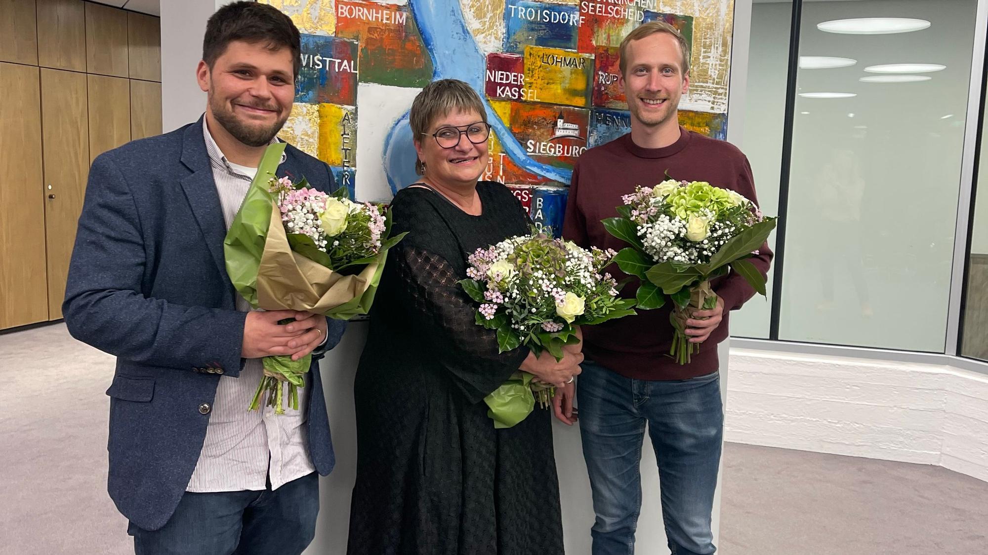 Zwei Männer und eine Frau mit Blumensträußen.