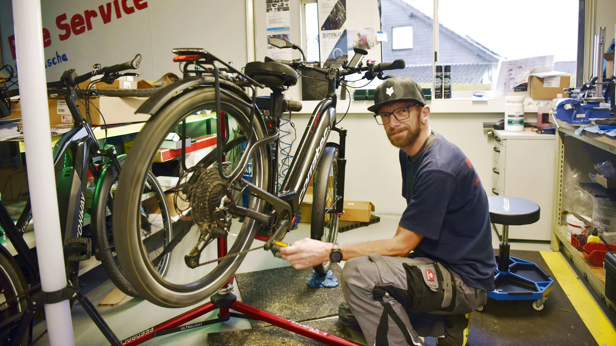 Sascha Tendler kniet in seiner Fahrradwerkstatt in Schöneseiffen vor einem Fahrrad und testet die Pedale. Er trägt Arbeitshose, ein dunkelblaues T-Shirt und eine Kappe.