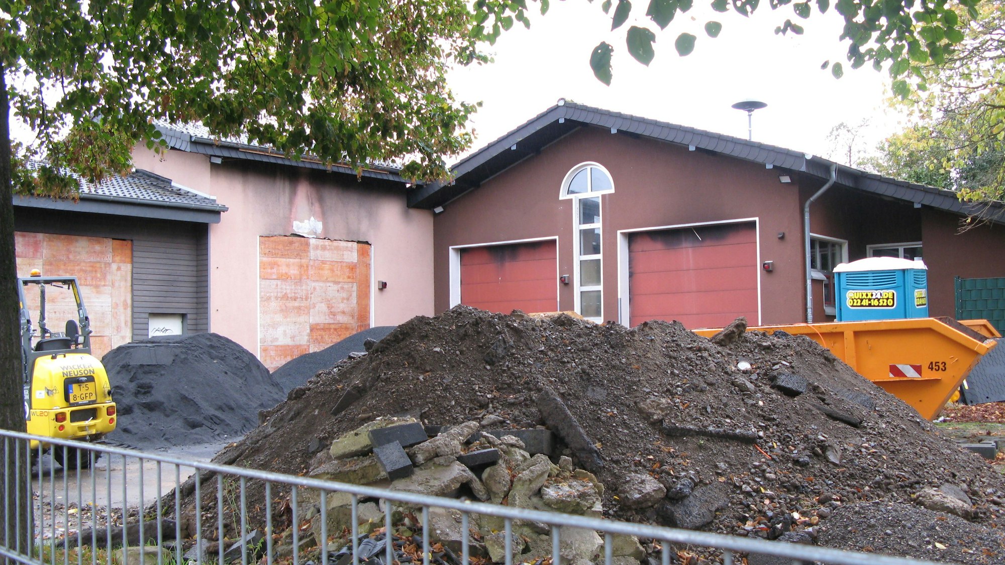 Vor einem eingeschossigen Gebäude türmt sich Schutt, ein kleiner Bagger, ein Container und ein Baustellenklo stehen dort.