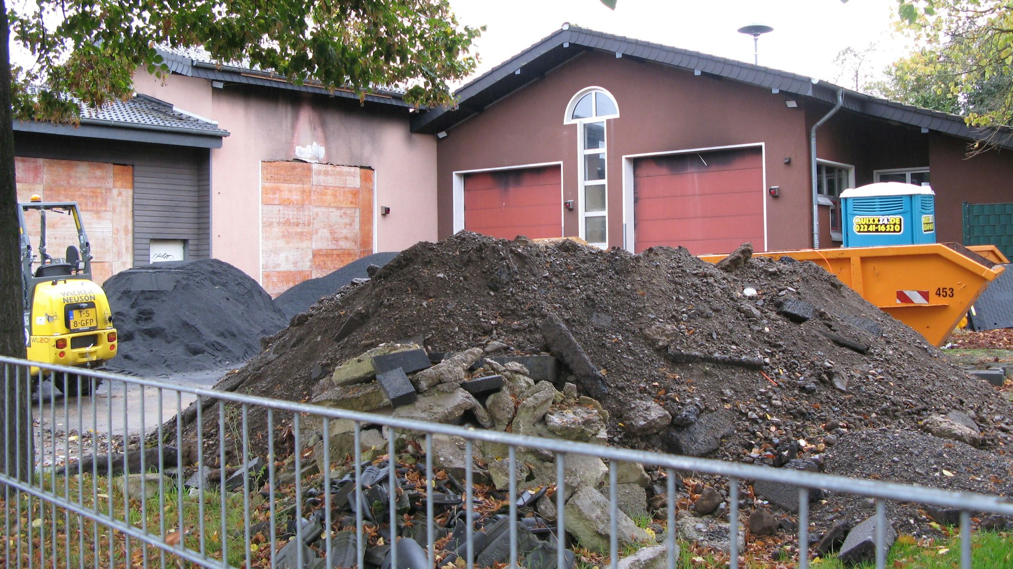 Ein Bauzaun, dahinter ein Abfallcontainer, ein Berg mit Abrissmaterial, ein kleiner Bagger und ein ehemaliges Feuerwehrhaus.