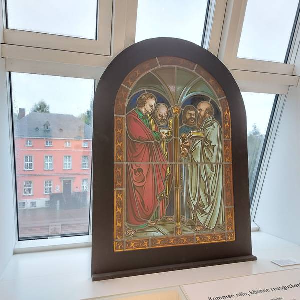 Ein historisches Buntglasfenster vor einem modernen Dachfenster; im Hintergrund ein historisches Herrenhaus mit Sprossenfenstern.