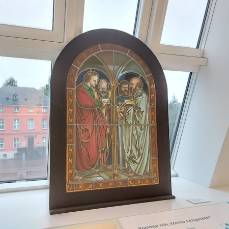 Ein historisches Buntglasfenster vor einem modernen Dachfenster; im Hintergrund ein historisches Herrenhaus mit Sprossenfenstern.