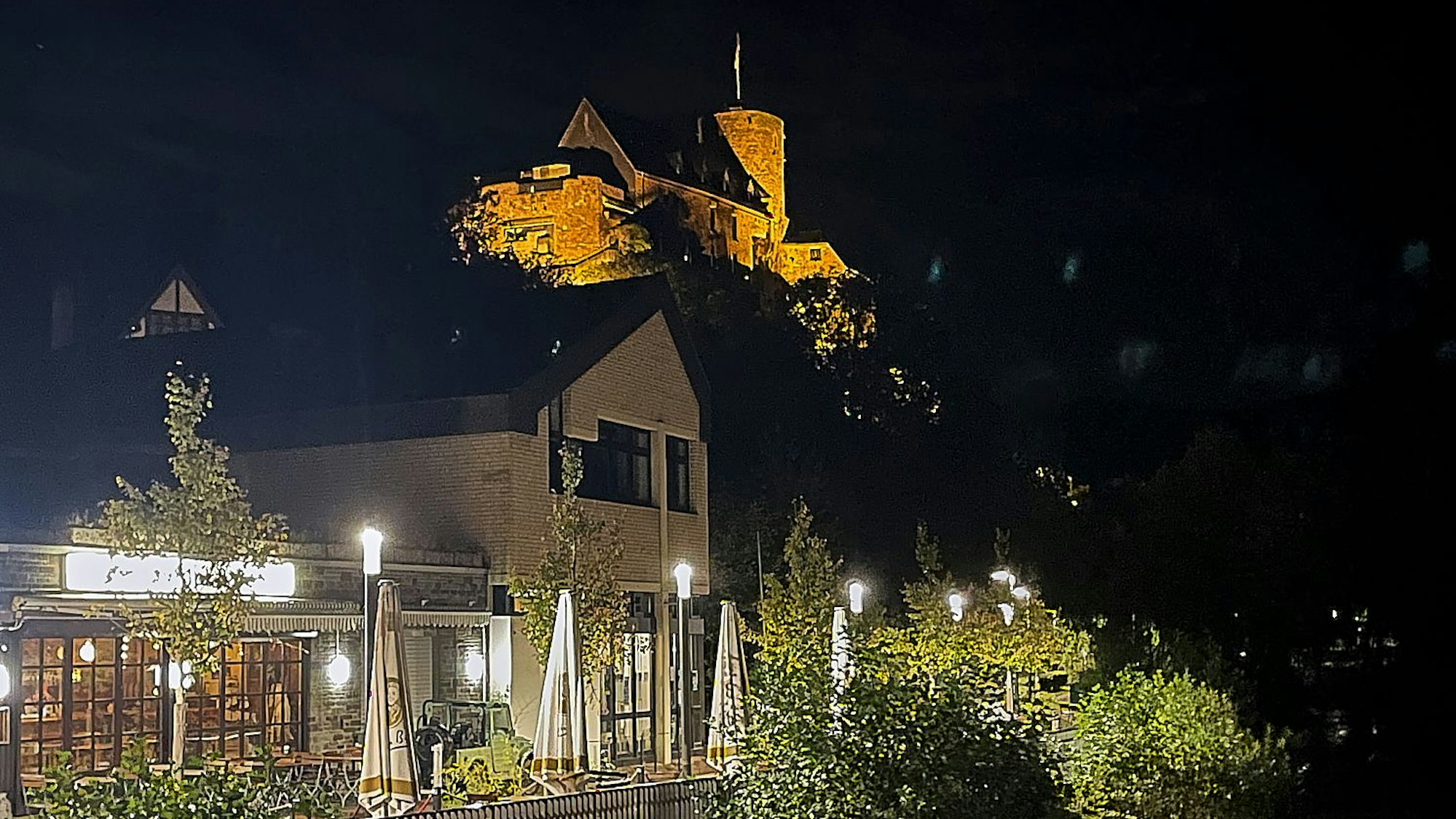 Das Bild zeigt die erleuchtete Heimbacher Burg im Dunklen. Im Vordergrund ist die Uferpromenade zu sehen.