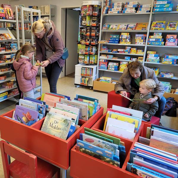 Eine Frau und ihre Tochter stöbern in den Regalen der Bücherei, eine Frau liest einem kleinen Jungen aus einem Buch vor.