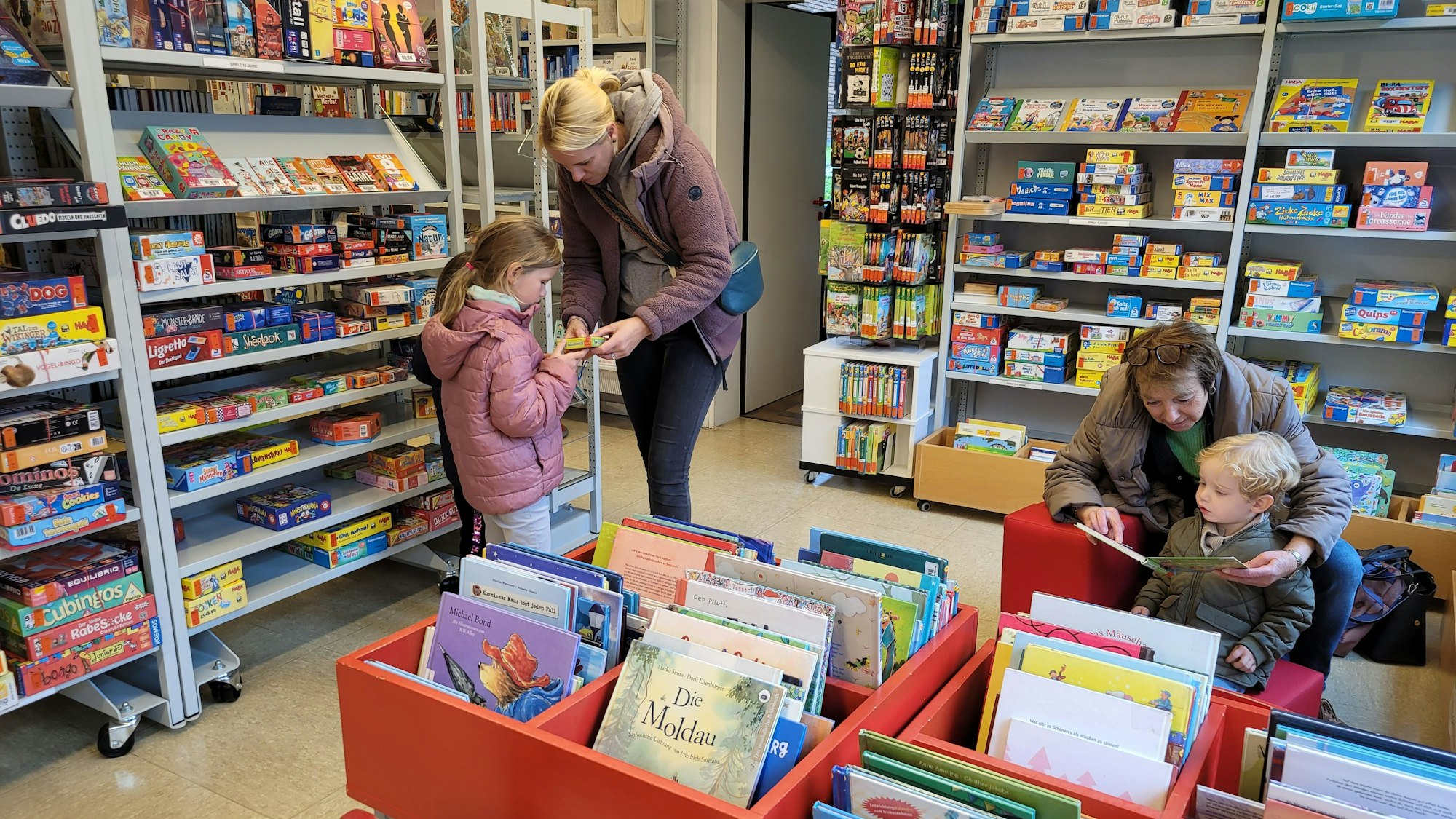 Eine Frau und ihre Tochter stöbern in den Regalen der Bücherei, eine Frau liest einem kleinen Jungen aus einem Buch vor.