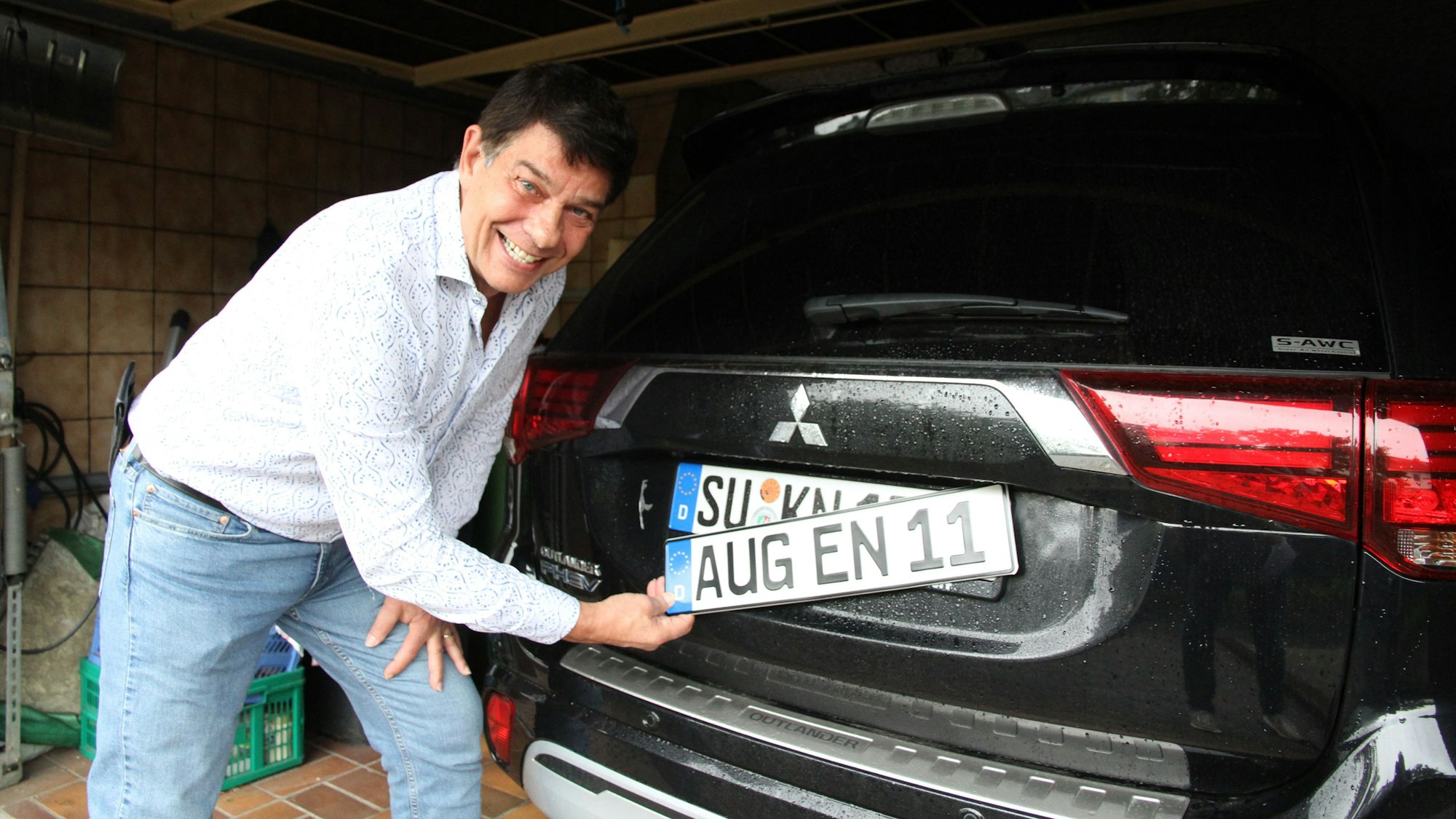 Ein Mann hält ein fiktives Kennzeichen vor das echte Nummernschild an der Hecktür seines Autos.