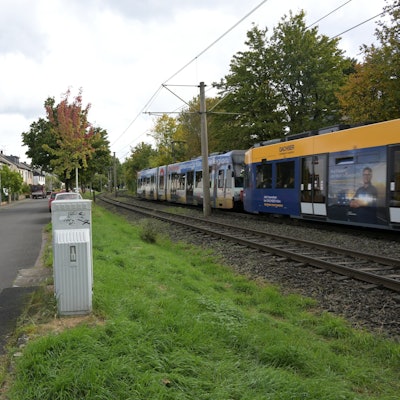 Links stehen Häuser, davor sind Hecken gepflanzt, in der Mitte ist die schmale Fahrbahn, rechts sieht man einen kleinen Grünstreifen, dann kommen die Bahngleise.
