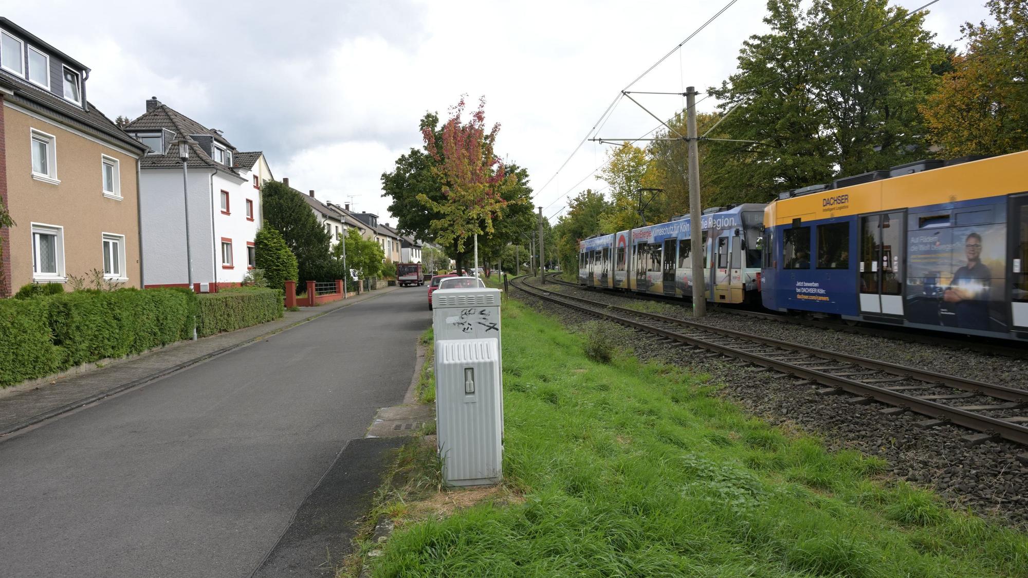 Links stehen Häuser, davor sind Hecken gepflanzt, in der Mitte ist die schmale Fahrbahn, rechts sieht man einen kleinen Grünstreifen, dann kommen die Bahngleise.