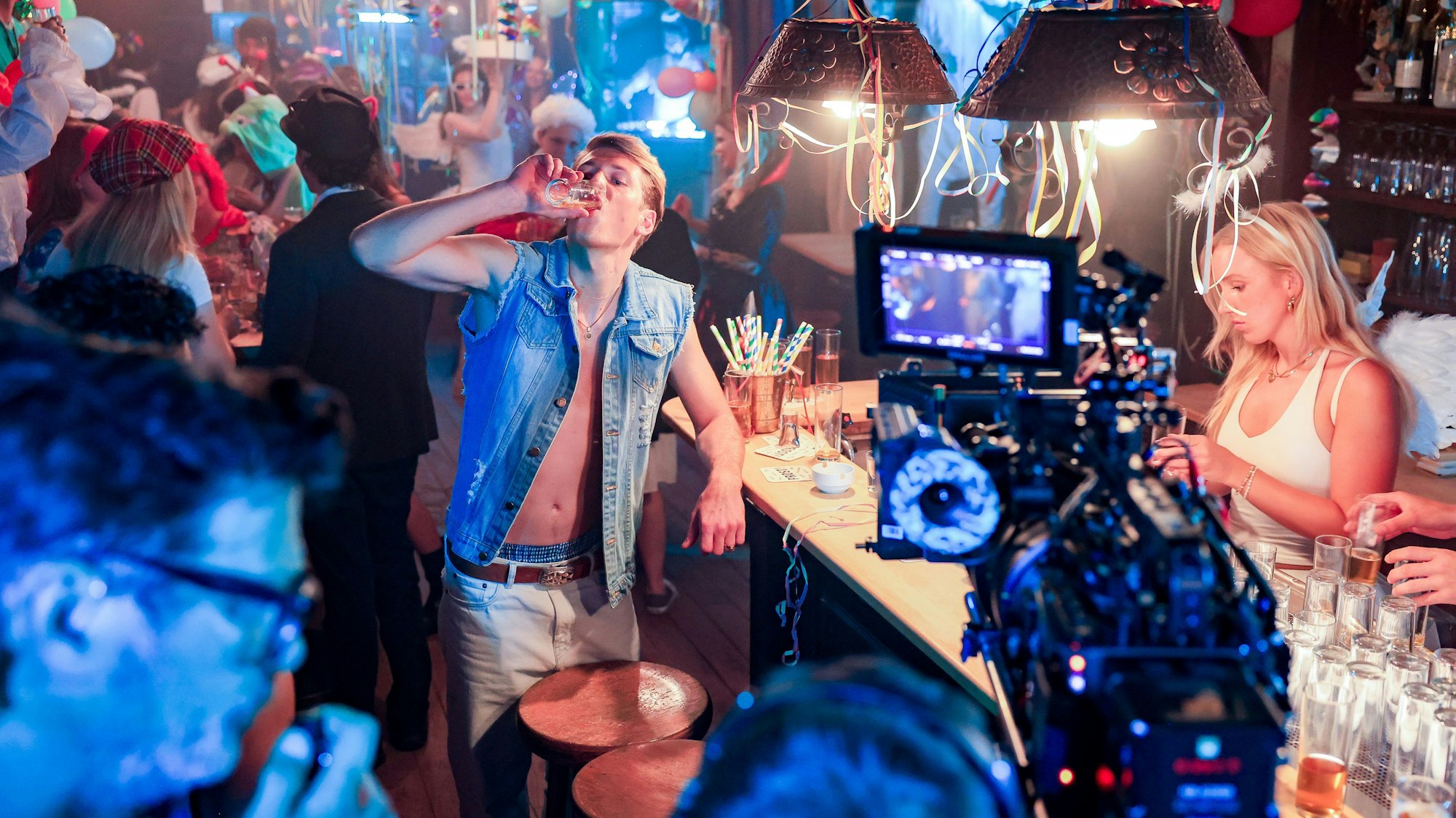 Blick in mit Konfetti geschmücktes Brauhaus voll verkleideter Menschen, vorne ist eine Filmkamera zu sehen.