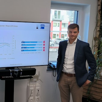 Zwischen zwei Männern steht ein großer Bildschirm, auf dem in Diagrammen die schrumpfende Wirtschaft angezeigt wird.