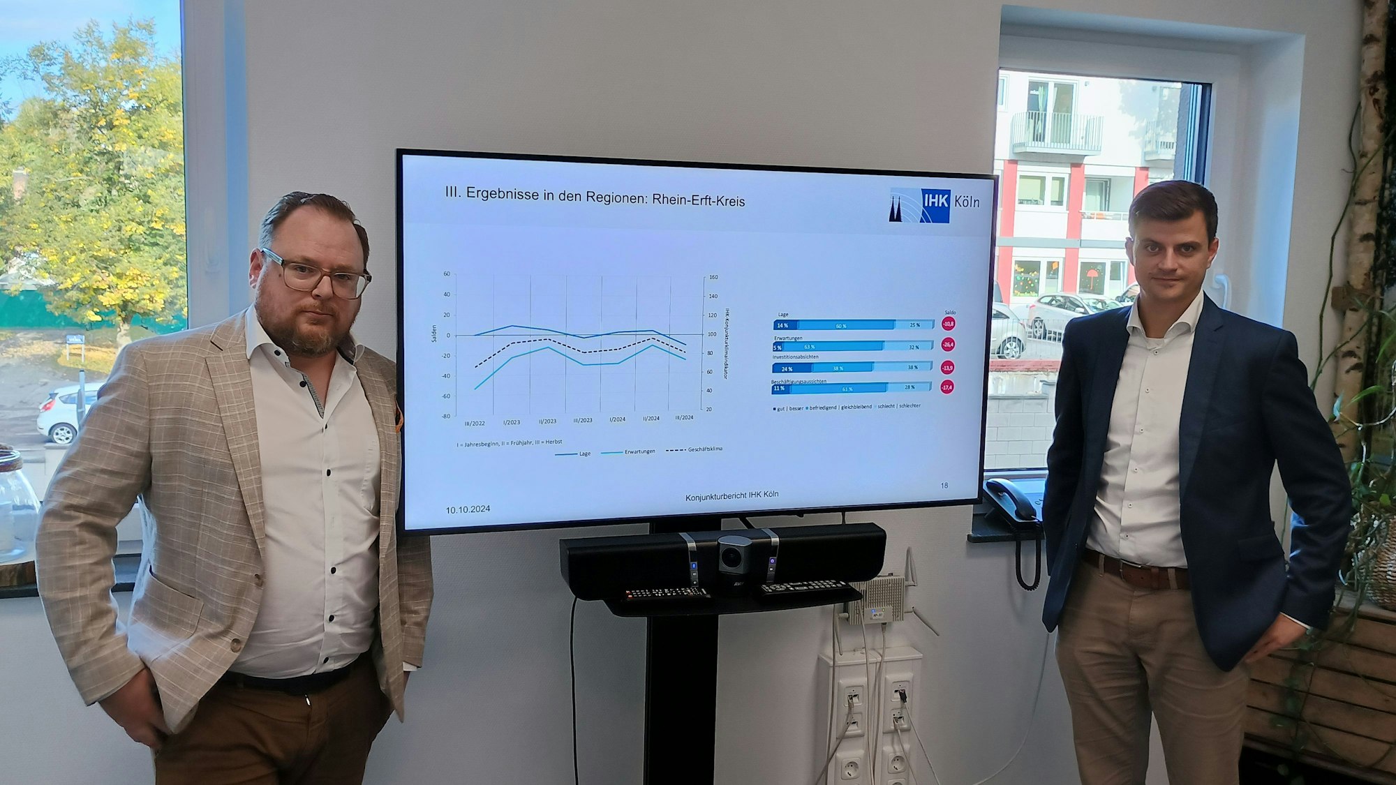 Zwischen zwei Männern steht ein großer Bildschirm, auf dem in Diagrammen die schrumpfende Wirtschaft angezeigt wird.