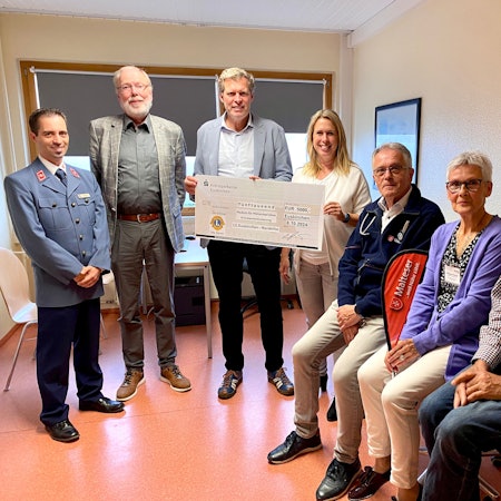 Sieben Menschen stehen und sitzen in einer Arztpraxis, zwei von ihnen halten einen symbolischen Scheck in den Händen.