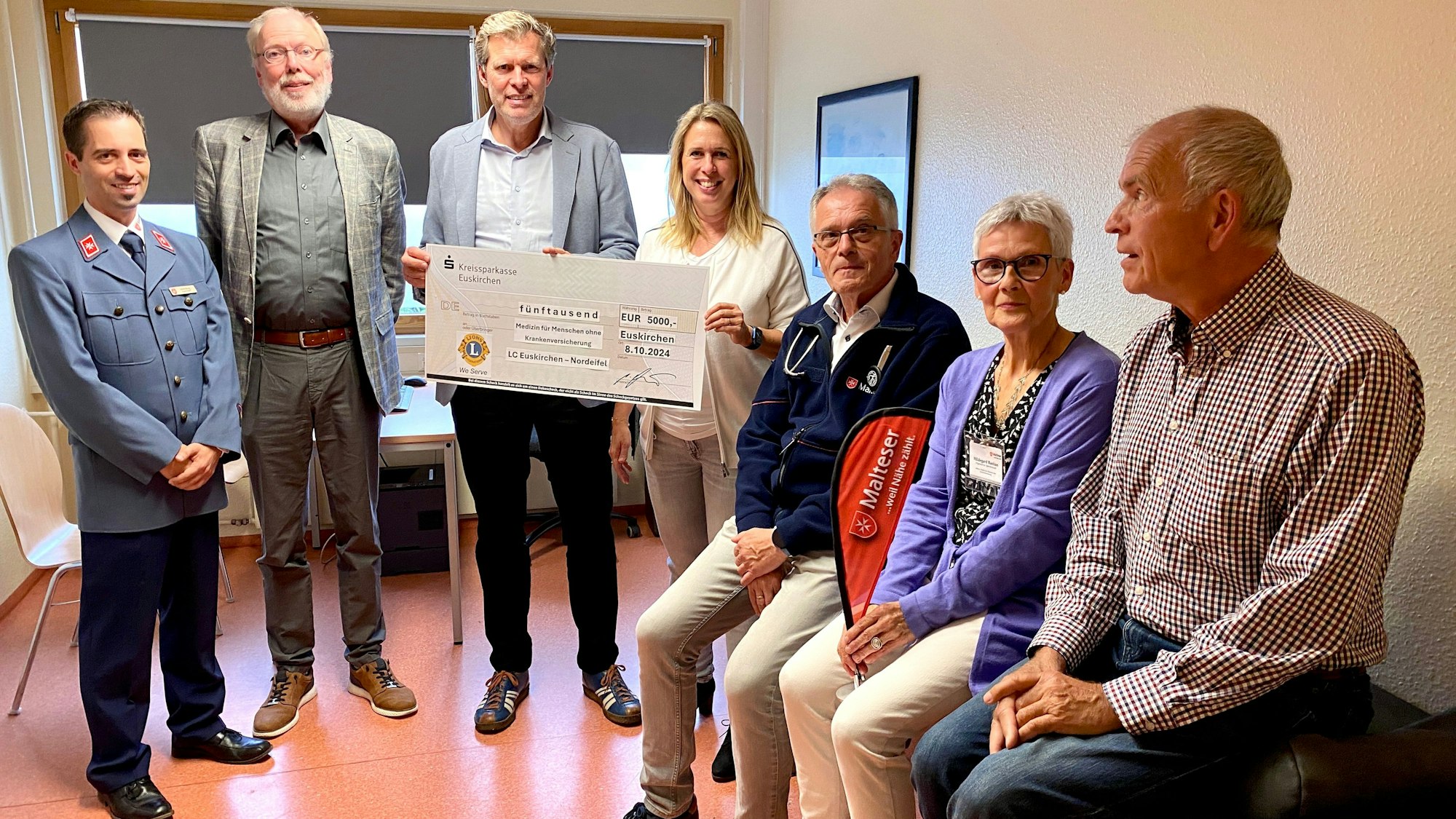 Sieben Menschen stehen und sitzen in einer Arztpraxis, zwei von ihnen halten einen symbolischen Scheck in den Händen.
