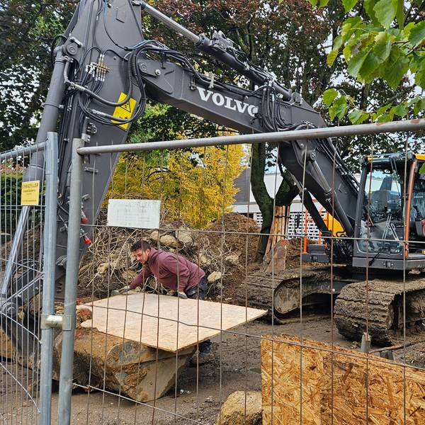 Ein Bagger steht hinter einem Bauzaun vor einer Schule.
