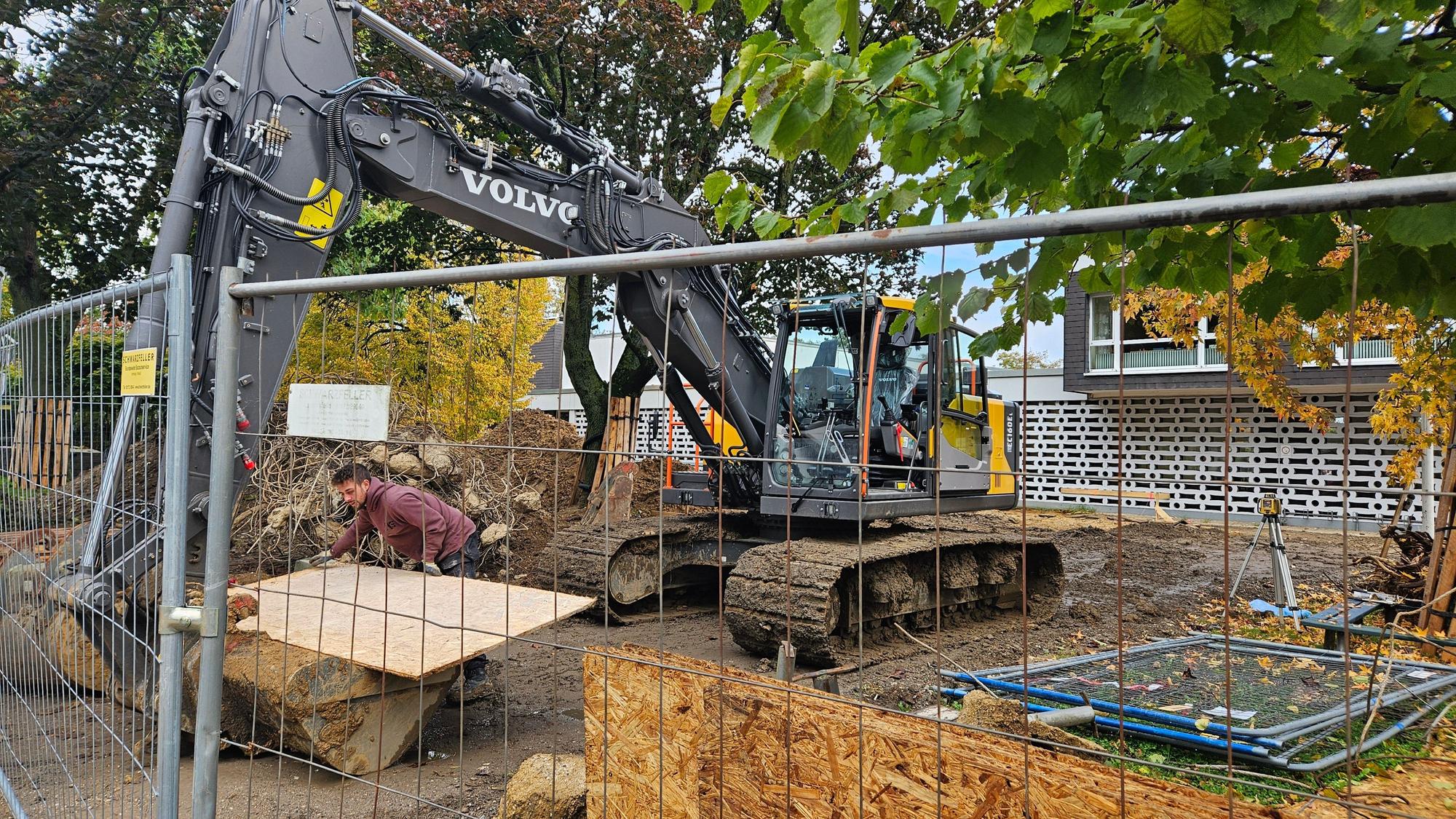 Ein Bagger steht hinter einem Bauzaun vor einer Schule.