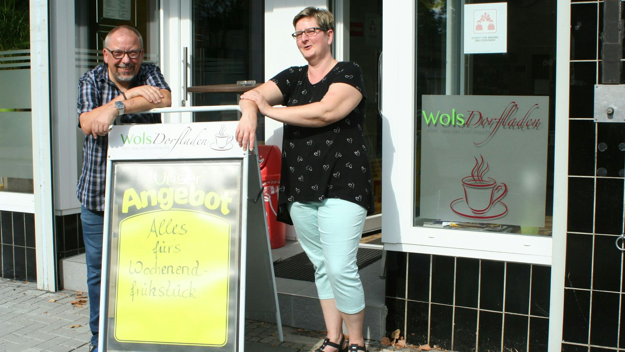 Ein Mann und eine Frau stehen vor einem Ladenlokal mit dem Logo „WolsDorfladen“.