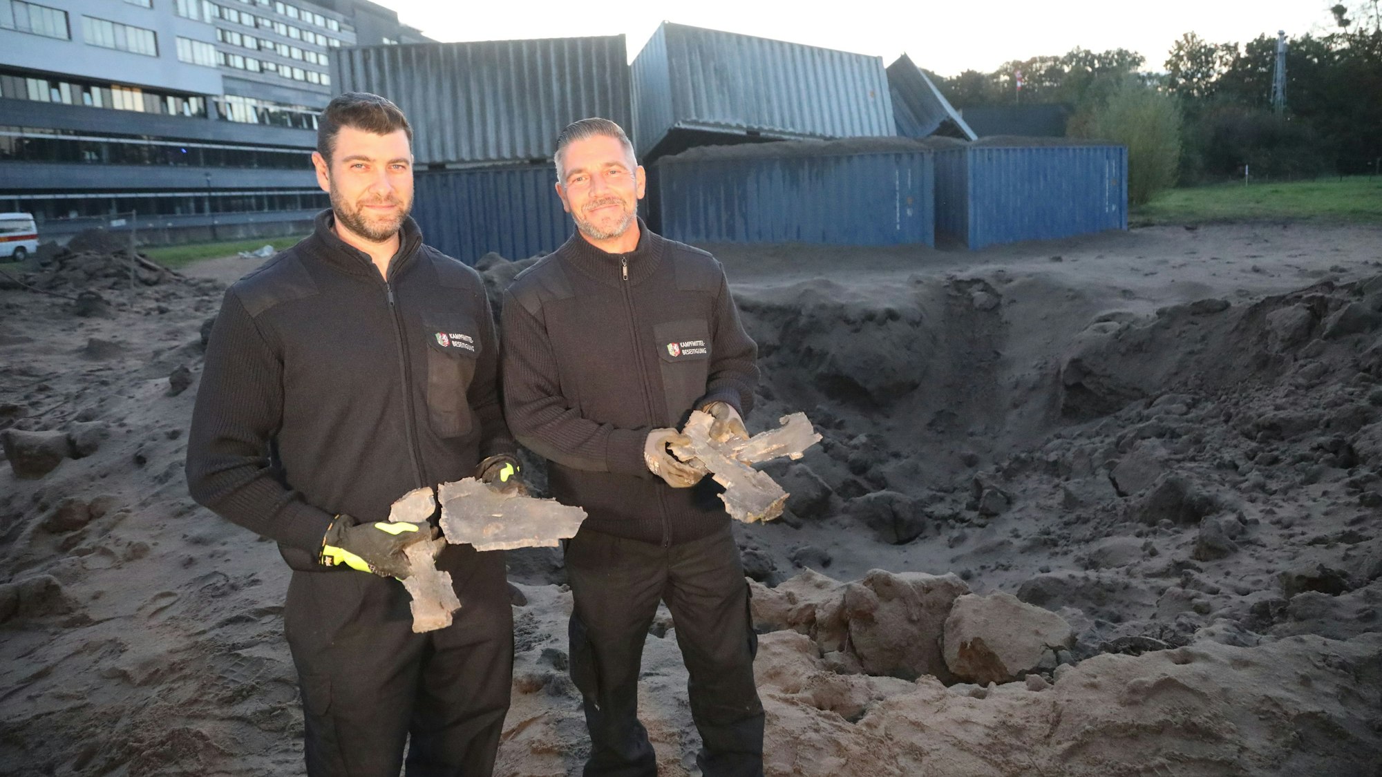 Marcel Biewald und Michael Hilgers (l.) vom Kampfmittelräumdienst halten die Reste der Bombe in der Hand. Im Hintergrund sind das Krankenhaus Merheim und ein Krater zu sehen.