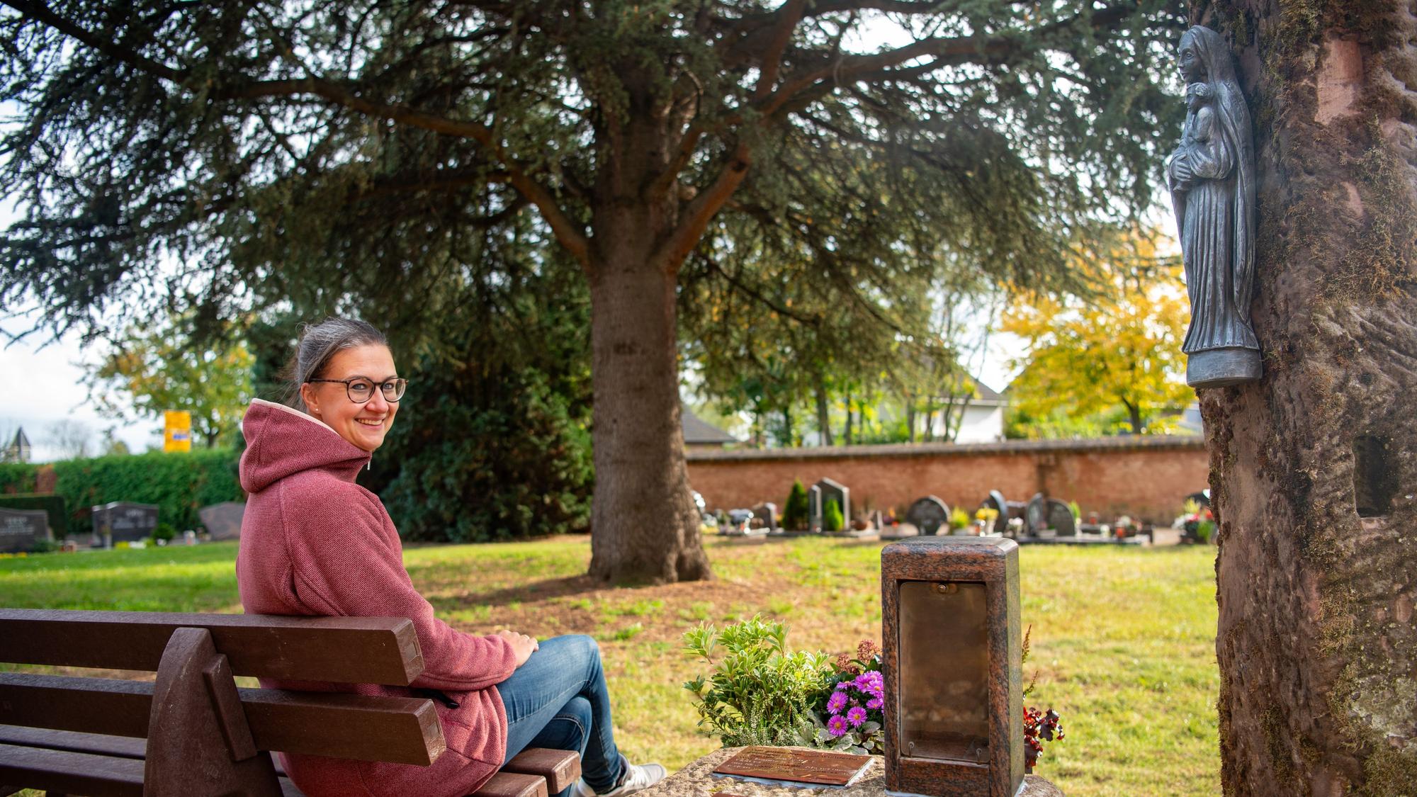 Krystina Antons sitzt auf einer Bank, rechts daneben steht ein Stein mit einem Blumengesteck und einer Halterung für ein Grablicht, rechts daneben steht eine Stele, an der eine Marienfigur hängt.