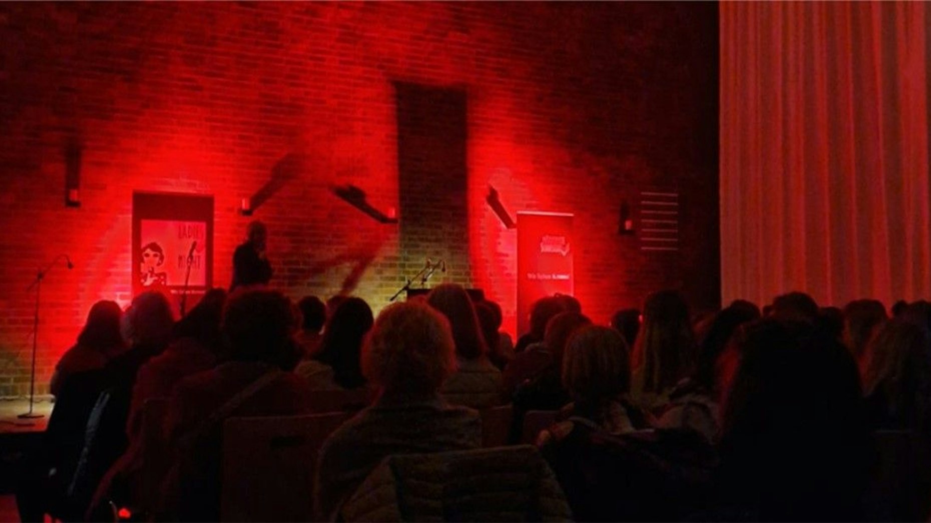 Der Saal mit Publikum ist in tiefrotes Licht gehüllt, hinten eine Bühne.